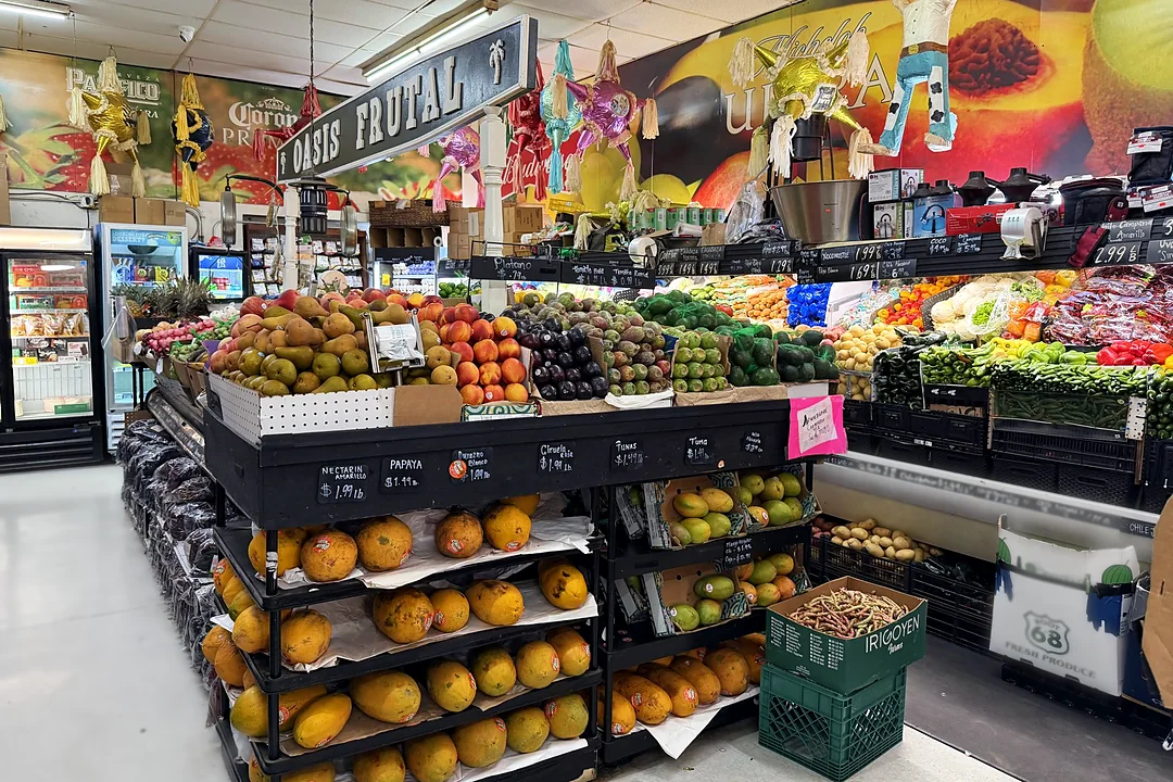 El pasillo de frutas y verduras bajo el letrero Oasis Frutal: papayas, mangos, nectarinas, aguacates y chiles, con piñatas colgando del techo